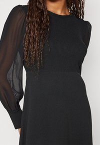 Schwarzes Kleid mit geripptem Stoff im Oberkörper und durchsichtigen Puffärmeln. Der Stoff wirkt glatt und fließend.