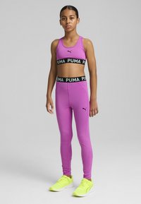 Lilla sports-bh og leggings sæt med sort "PUMA" taljebånd, der har et lille sort logo. Klar gule atletiske sko fuldender outfittet.