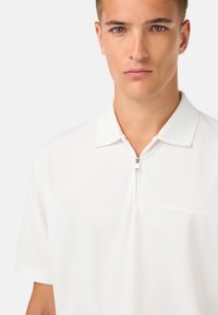 Bílá polo tričko s límcem na zip. Krátké rukávy, hladká textura a jemná náprsní kapsa. Přiléhavý střih a čistý design.