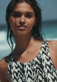 Jauna moteris su šlapiais plaukais, dėvinti juodai baltą raštuotą berankovį viršų, stovinti lauke šalia vandenyno saulėta diena.