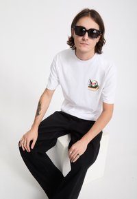 Jeune homme aux cheveux mi-longs et lunettes de soleil portant un t-shirt blanc avec un motif de crâne, un pantalon noir et des bagues argentées, assis sur un bloc blanc.