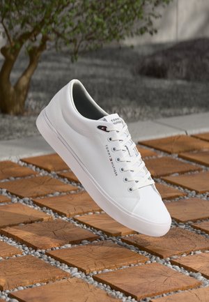 Baskets blanches Tommy Hilfiger avec lacets flottant au-dessus de dalles en bois avec des graviers entre elles en extérieur.