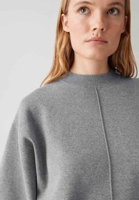 Junge Frau mit heller Haut und langen blonden Haaren, die einen grauen Strickpullover mit hohem Ausschnitt vor einem schlichten Hintergrund trägt.
