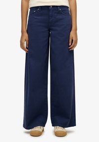 Pantalones de mezclilla de pierna ancha en azul marino con diseño de cinco bolsillos, botones frontales y una ligera textura. Combinados con zapatos de color claro.