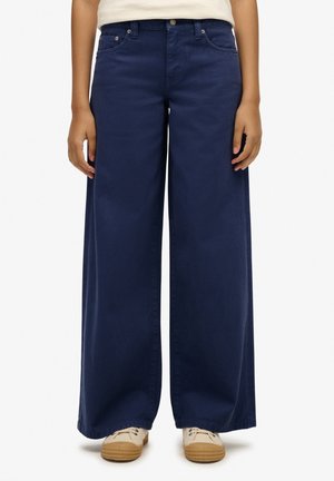 Bredde ben, marineblå denim bukser med fem lomme-design, frontknapper og let tekstur. Parret med lyse sko.