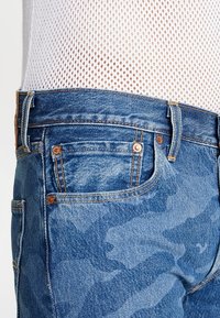 Vaaleansiniset farkut, joissa on haalistunut maastokuvio, viisi taskua, kuparinappulat ja ompeluyksityiskohtia vyötäröllä ja taskuissa.