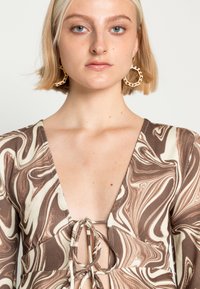 Roupão de manga longa com um decote em V profundo, padrão de mármore castanho e creme, textura canelada e um detalhe de laço no peito. Brincos de argola dourados.