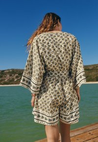 Combinaison légère beige avec motif de feuillage noir, à manches larges et un bandeau assorti le long de l'ourlet, portée devant un paysage côtier.