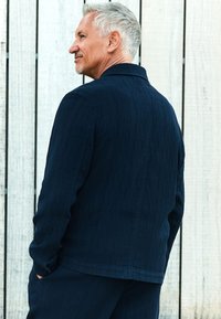 Marineblå tekstureret jakke med lange ærmer og krave, med sidelommer og en lige kant, båret over matchende bukser.