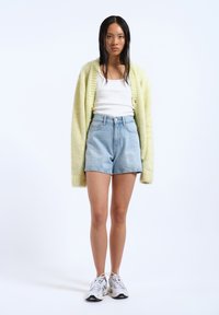 Un cardigan tricoté jaune clair sur un débardeur blanc, associé à un short en denim bleu clair taille haute et des chaussures de sport blanches.