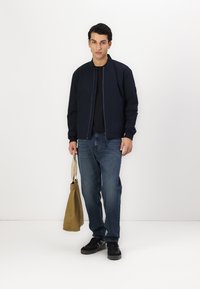 Giacca bomber blu navy con zip, t-shirt nera, jeans blu e sneakers nere. Tiene una borsa tote marrone chiaro. Sfondo bianco semplice.