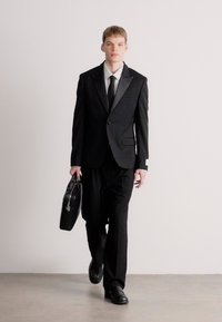 Μαύρο κοστούμι με σατέν πέτο, λευκό πουκάμισο και μαύρη γραβάτα. Το μοντέλο κρατά μια δερμάτινη τσάντα και φοράει μαύρα παπούτσια. Απλός, κομψός σχεδιασμός.