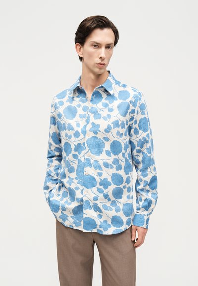 Άνδρας που φοράει λευκό πουκάμισο με μπλε φλοράλ σχέδιο και ανοιχτό καφέ παντελόνι, στέκεται με τα χέρια στις τσέπες μπροστά σε απλό φόντο.