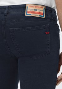 Jeans en denim bleu foncé vue arrière avec une étiquette rectangulaire Diesel Industry au-dessus de la poche droite et un logo rouge brodé dans le coin de la poche.