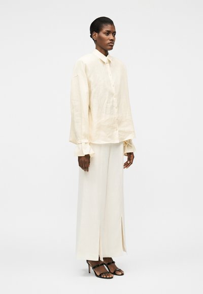 Γυναίκα που στέκεται φορώντας χαλαρό εκρού πουκάμισο με κουμπιά και βολάν στα μανίκια, παντελόνι με φαρδιά μπατζάκια και μπροστινές σχισμές, και μαύρα σανδάλια με τακούνι.
