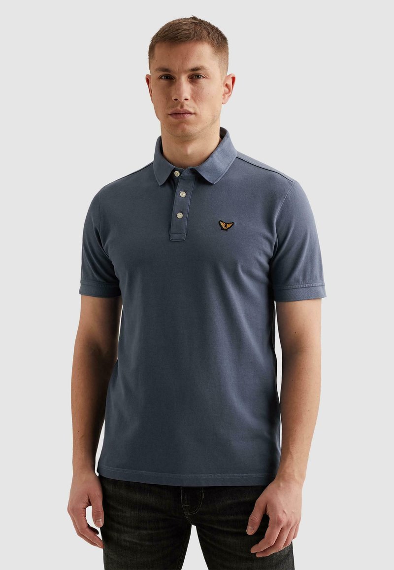 Ung mand iført en blå poloshirt med korte ærmer og et broderet gult og sort fuglelogo på venstre bryst.