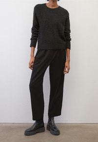 Pull en tricot gris foncé avec un col rond, associé à un pantalon noir ample et des bottines noires épaisses sur un fond neutre.