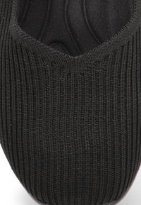 Zapato de punto negro con punta redondeada y textura acanalada. Diseño flexible sin costuras ni herrajes visibles. Estética suave y minimalista.