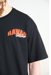 Černé tričko nošené osobou, s červeným textem "HAWAII" a bílým kurzívním "aloha" na hrudi.