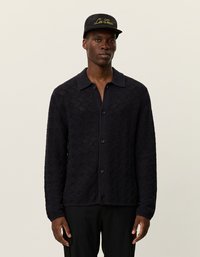 Egy férfi fekete, textúrált gombos kardigánt, fekete nadrágot és fekete sapkát visel, amelyen arany színű "Les Deux" hímzés található, semleges háttér előtt áll.