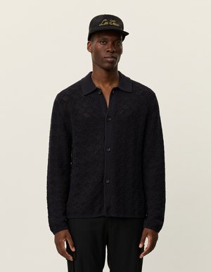 Mann trägt eine schwarze strukturierte Knopfleiste-Cardigan, schwarze Hose und eine schwarze Mütze mit goldener "Les Deux"-Stickerei, steht vor einem neutralen Hintergrund.
