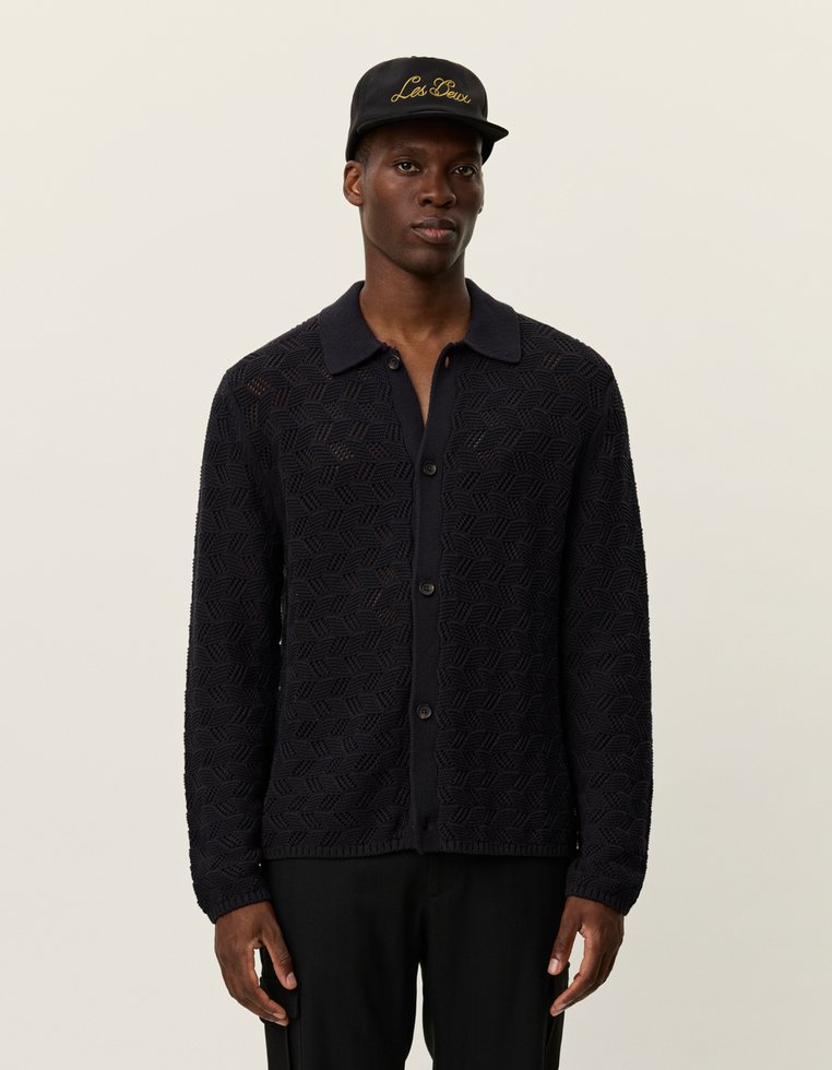 Egy férfi fekete, textúrált gombos kardigánt, fekete nadrágot és fekete sapkát visel, amelyen arany színű "Les Deux" hímzés található, semleges háttér előtt áll.