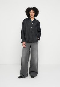 Πουκάμισο από τζιν με κουμπιά σε σκούρο μπλε χρώμα και μακριά μανίκια, συνδυασμένο με γκρι τζιν παντελόνι φαρδιάς γραμμής με ξεθωριασμένη υφή και ορατές ραφές.