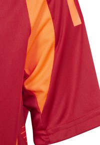Maglietta sportiva rossa con un pannello laterale arancione. Il tessuto è liscio con una texture in rete traspirante. Presenta maniche corte e un orlo dritto.