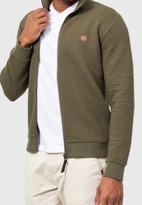 Veste zippée vert olive avec un motif texturé, poignets et col côtelés. Dispose d'un petit patch logo sur la poitrine. Portée par-dessus une chemise blanche.