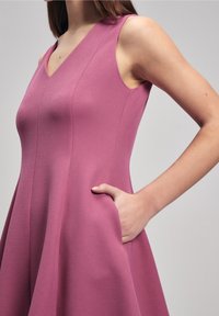 Robe ajustée sans manche rose avec un décolleté en V, en tissu texturé, jupe évasée et poches latérales, mettant en avant des lignes épurées et une silhouette structurée.