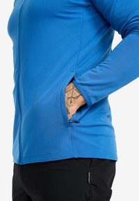 Blå långärmad skjorta i slätt, elastiskt tyg med sidfickor, dragkedjestängning och kontrasterande svarta byxor.