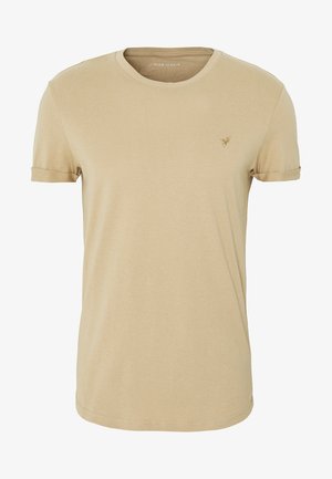 Camiseta de algodón beige con cuello redondo, mangas cortas y puños doblados. Presenta un pequeño logotipo bordado en el pecho. Textura suave.
