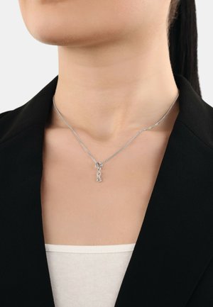 Silberne Halskette mit einem Anhänger in Form des Unendlichkeitssymbols, ausgestattet mit einer schlanken, glatten Textur und einer feinen Kette, die über einem weißen Oberteil und einem schwarzen Blazer getragen wird.
