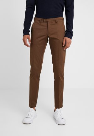 Mand iført slim-fit brune bukser, hvide sneakers og en mørk langærmet top står mod en ensfarvet lys baggrund.