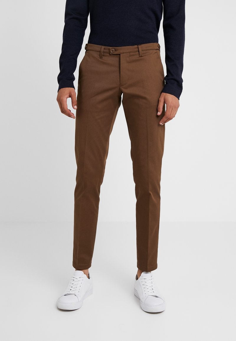 Homme portant un pantalon slim marron, des baskets blanches et un haut à manches longues foncé, debout devant un fond clair uni.
