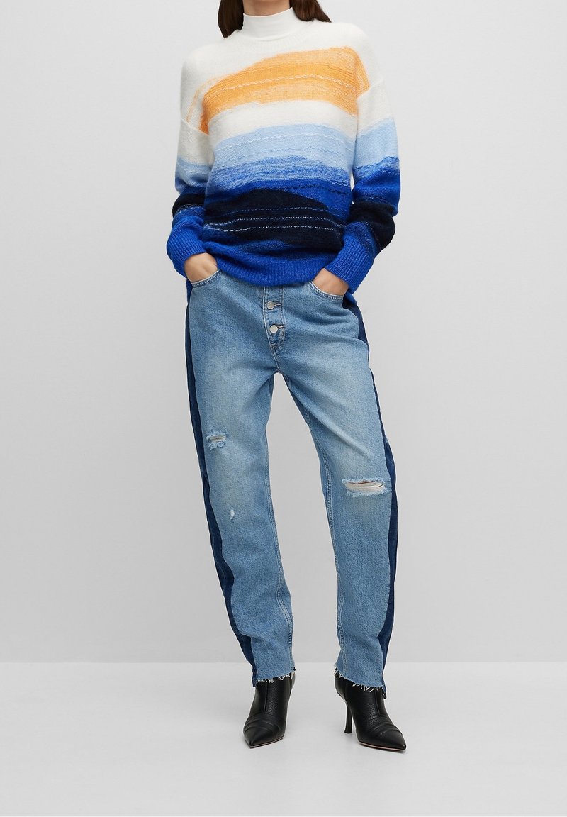 Sweter dzianinowy w poziome paski w kolorze pomarańczowym, niebieskim i białym, zestawiony z jasnoniebieskimi d�żinsami z bocznym granatowym paskiem i przetarciami.