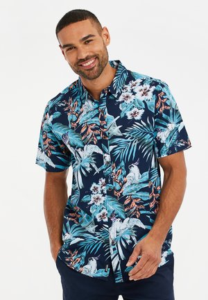 Chemise à manches courtes avec un fond bleu marine ornée de motifs tropicaux de fleurs et d'oiseaux en turquoise, rose et blanc.