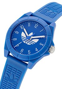 Montre-bracelet en silicone bleu avec un cadran rond, marquages horaires blancs et trois aiguilles. Présente un logo en relief sur le bracelet et un bandeau texturé.