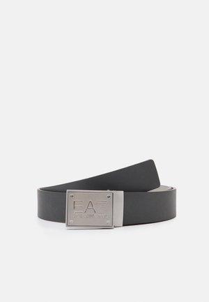 Ceinture en cuir noir avec boucle rectangulaire en métal argenté gravée du logo « EA7 Emporio Armani », enroulée sur une surface blanche.
