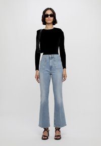 Svart langermet topp, høythengende lyseblå flagrende jeans, svarte stroppesandal, og liten svart skulderveske. Glatte teksturer og moderne design.