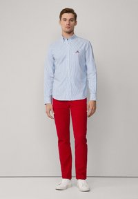 Camisa de botones a rayas azul y blanca, combinada con pantalones rojos y zapatillas blancas. La camisa presenta un pequeño logo bordado.