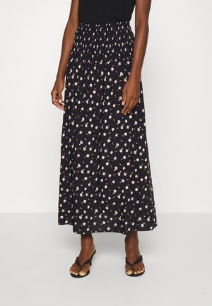 Femme portant une jupe noire à fleurs longueur cheville avec une taille smockée et des sandales à talons noires, debout devant un fond blanc.