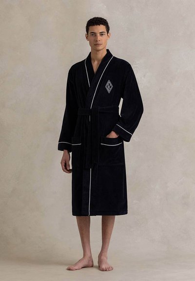 Μαύρη, βελούδινη ρόμπα μέχρι το γόνατο με ζώνη στη μέση, λευκή διακόσμηση και λογότυπο σε σχήμα ρόμβου. Διαθέτει δύο εμπρόσθιες τσέπες και μακριά μανίκια.