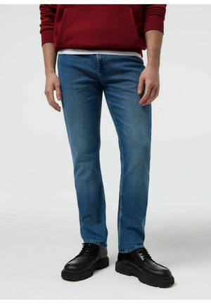 Mężczyzna w niebieskich obcisłych dżinsach, czarnych masywnych sznurowanych butach i czerwonej bluzie na jednolitym tle.