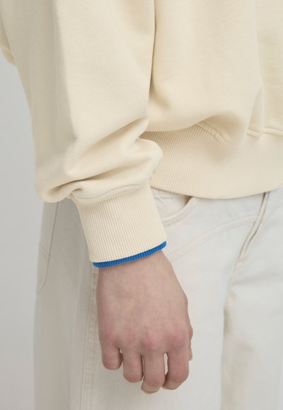 Sudadera color crema con puños acanalados que presentan un acento azul. La tela parece suave con un diseño de corte holgado.