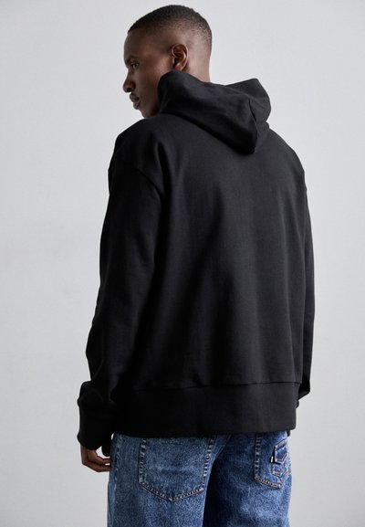 Μαύρο φούτερ με κουκούλα σε χαλαρή γραμμή, με ακατέργαστο τελείωμα και μαλακή υφή, συνδυασμένο με ανοιχτό μπλε τζιν παντελόνι με φθορές.