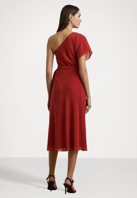 Punainen pilkullinen mekko yhdellä olalla, lentävä kangas, keskipitkä pituus, ja sidottu vyötäröysitys. Korkokengät täydentävät asua.