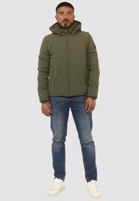 Giacca imbottita verde oliva con cappuccio, cerniera frontale e tasche laterali, abbinata a jeans blu e sneaker bianche. Design e texture chiari.