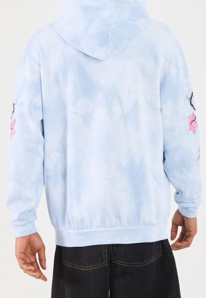 Lyseblå tie-dye hættetrøje med hætte, lyserød blomsterbroderi på ærmerne, båret af person i sorte bukser, vist bagfra og fra livet og op.