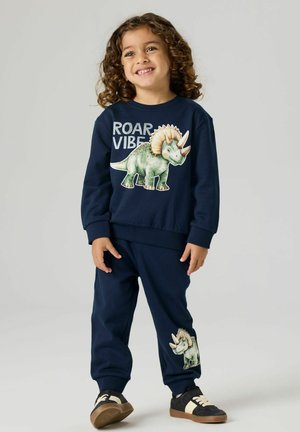 Smilende barn med krøllet hår iført navyblå sweatshirt og bukser med et grønt triceratops-dinosaurmotiv og teksten "ROAR VIBE".